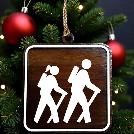 Wood hiking trail sign ornament showing female and male hikers with walking sticks, designed on dark walnut with white MDF overlay, hanging on a Christmas tree with red holiday ornaments in the background.