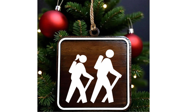 Wood hiking trail sign ornament showing female and male hikers with walking sticks, designed on dark walnut with white MDF overlay, hanging on a Christmas tree with red holiday ornaments in the background.