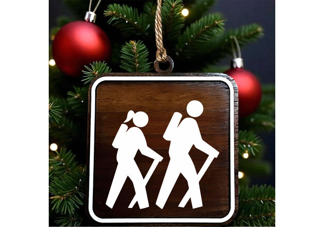 Wood hiking trail sign ornament showing female and male hikers with walking sticks, designed on dark walnut with white MDF overlay, hanging on a Christmas tree with red holiday ornaments in the background.