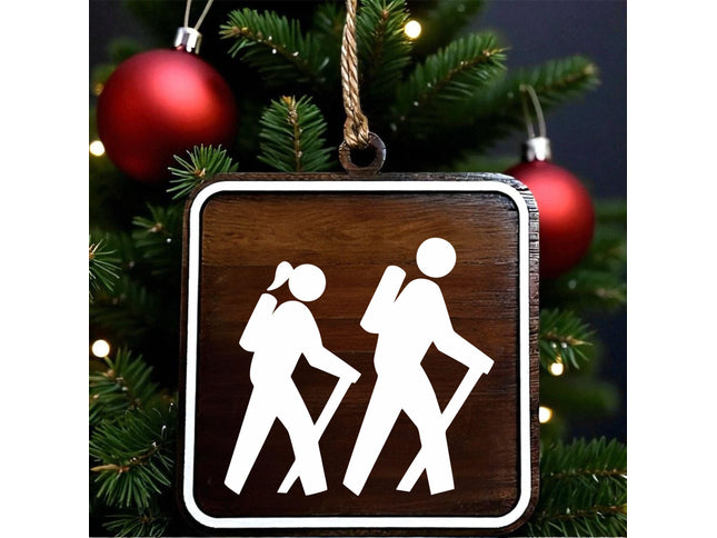 Wood hiking trail sign ornament showing female and male hikers with walking sticks, designed on dark walnut with white MDF overlay, hanging on a Christmas tree with red holiday ornaments in the background.