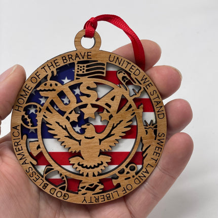 A wooden ornament with a red, white, and blue American flag design. The ornament is shaped like a circle and has the words &#39;God Bless America&#39;, &#39;Home of the Brave&#39;, &#39;United We Stand&#39;, &#39;Sweet Land of Liberty&#39; written around the edge. The center of the ornament features a bald eagle with the word &#39;USA&#39; in the background.