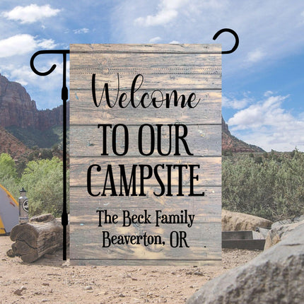 Custom “Welcome to Our Campsite” garden flag displayed at a campsite with mountains and trees in the background. Features a rustic wood-look design and personalized text reading “The Beck Family, Beaverton, OR.” Shown hanging on a black garden flag stand.