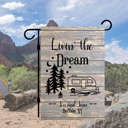 Custom “Livin’ the Dream” camper garden flag displayed at a scenic campsite with mountains and desert landscape in the background. Features a rustic wood-look design, pine trees, stars, a camper graphic, and personalized text reading “Jen and Jenn, Buffalo, NY,” hung on a black garden flag stand.