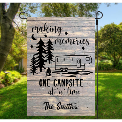 Custom “Making Memories One Campsite at a Time” garden flag displayed outdoors in a backyard setting. Features a rustic wood-look background, pine trees, stars, a camper graphic, and personalized text reading “The Smith’s.” Hung on a black metal garden flag stand.