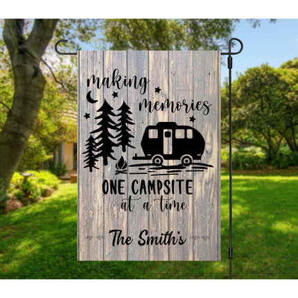 Custom “Making Memories One Campsite at a Time” garden flag displayed in a backyard setting. Features a rustic wood-look background, pine trees, stars, a small rounded camper graphic, and personalized text reading “The Smith’s.” Hung on a black metal garden flag stand.