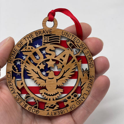 Close-up of a patriotic wooden eagle ornament held in hand, showing detailed laser-cut design. Features an American flag background, eagle centerpiece, and engraved border text including “Home of the Brave,” “United We Stand,” and “God Bless America.” Red ribbon attached for hanging.