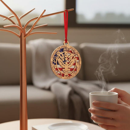 Patriotic wooden eagle ornament hanging from a modern metal display stand. Features a laser-cut design with an American flag background and engraved border text. A hand holding a warm cup is visible nearby, creating a cozy home setting. Great for gifting or year-round display.