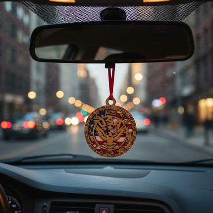 Patriotic wooden eagle ornament hanging from a car’s rearview mirror with a red ribbon. The ornament features a layered American flag background and eagle cutout with engraved text around the border. City street and evening lights are softly blurred in the background.
