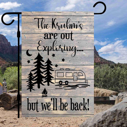 Custom “The Kruhm’s Are Out Exploring… but we’ll be back!” garden flag displayed at a scenic campsite with mountains and desert landscape in the background. Features a rustic wood-look design, pine trees, stars, a camper graphic, and personalized family name. Hung on a black garden flag stand.