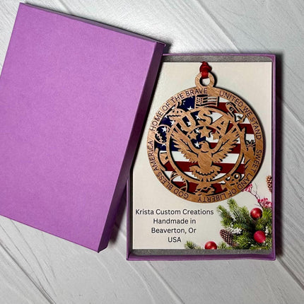 Patriotic wooden eagle ornament displayed in a purple gift box. The ornament features a layered American flag background with an eagle cutout and text around the border including “Home of the Brave,” “United We Stand,” and “God Bless America.” Card inside reads “Krista Custom Creations, Handmade in Beaverton, OR, USA.”