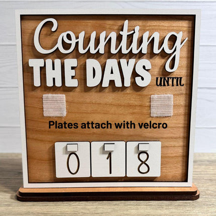 Interchangeable wooden countdown sign with “Counting The Days Until” text and rustic wood background, showing two Velcro attachment squares for interchangeable plates and number tiles displaying 018 on a tabletop stand.