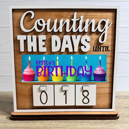 Wooden interchangeable countdown sign displaying “Counting The Days Until” with Birthday plate featuring colorful cupcakes and candles, and number tiles showing 018 on a tabletop stand.