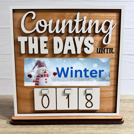 Wooden interchangeable countdown sign displaying “Counting The Days Until” with Winter plate featuring a snowman design and number tiles showing 018 on a tabletop stand.