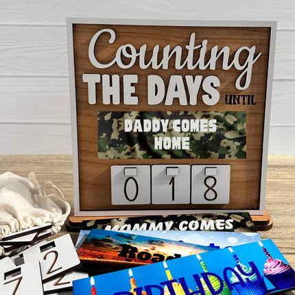 Wooden countdown sign reading “Counting the Days Until Daddy Comes Home,” featuring interchangeable number tiles displaying 018 days. The sign includes a camouflage-themed message plate and is shown with additional interchangeable plates and number tiles laid out in front, illustrating customizable countdown options for military homecomings and special events.