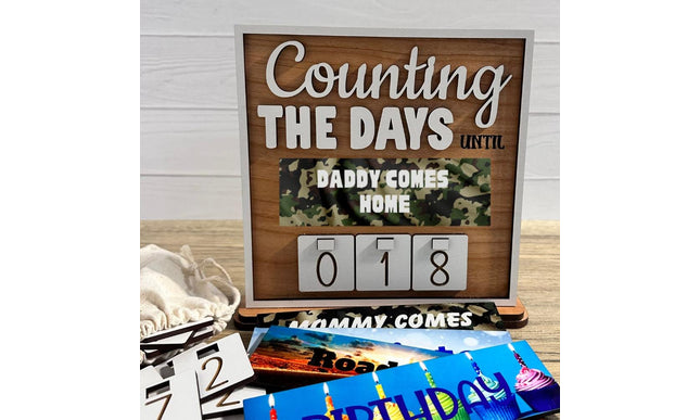 Wooden countdown sign reading “Counting the Days Until Daddy Comes Home,” featuring interchangeable number tiles displaying 018 days. The sign includes a camouflage-themed message plate and is shown with additional interchangeable plates and number tiles laid out in front, illustrating customizable countdown options for military homecomings and special events.